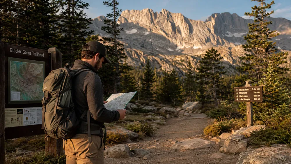 A hiker with a backpack reads a map at the start of a trail, surrounded by pine trees and mountains. Trail signs point the way on either side.