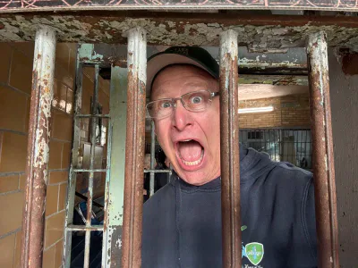 Shea wearing glasses and a cap yells with a surprised expression while standing behind rusty metal jail bars indoors.